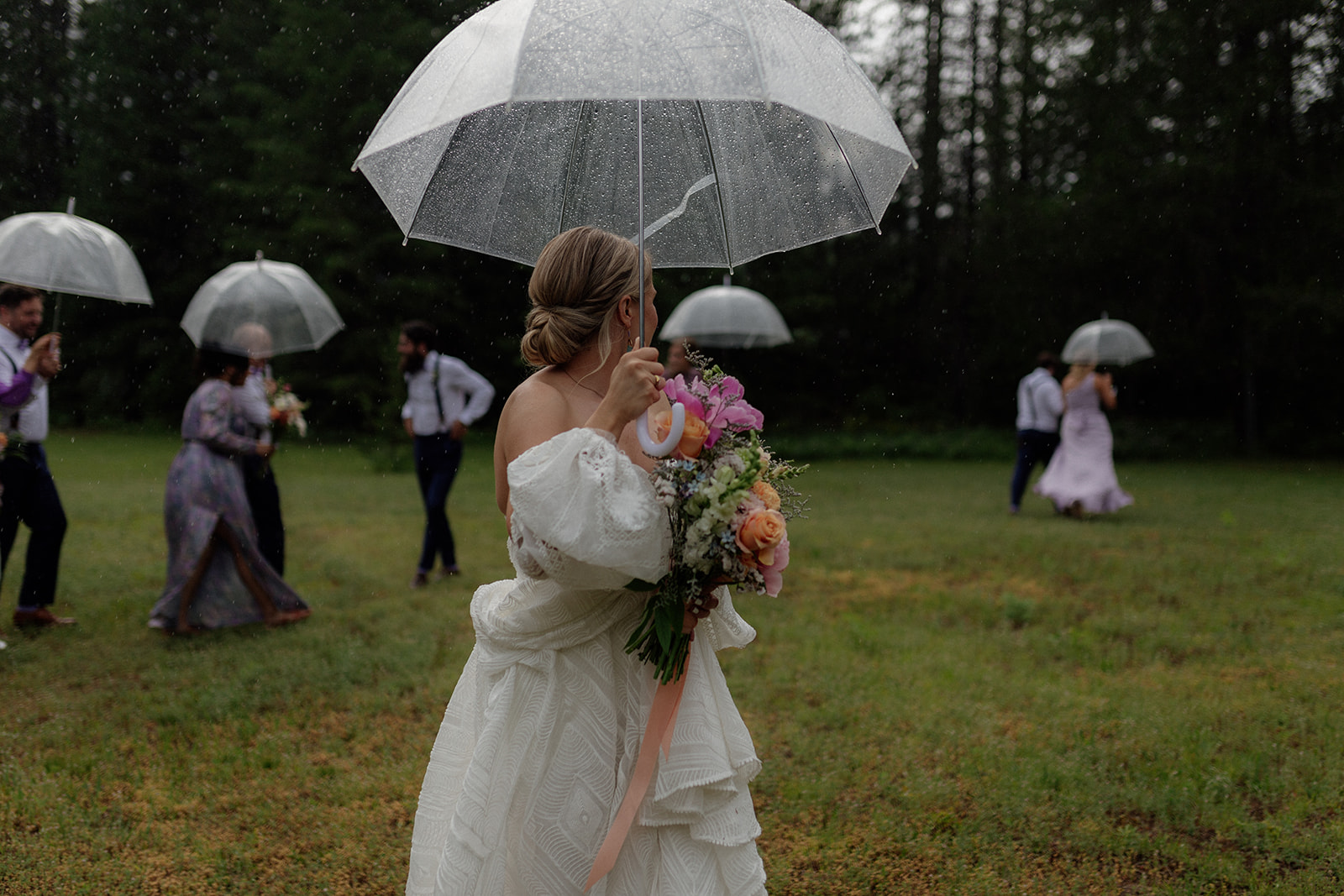 A bride in a beautiful white gown walks through a grassy field, holding a vibrant bouquet of flowers. It’s raining, and she and her wedding party are using clear umbrellas to shield themselves from the drizzle. The soft, overcast light and misty forest in the background create a romantic and serene atmosphere, capturing the unique charm of a Kootenay outdoor wedding, even in less-than-perfect weather.