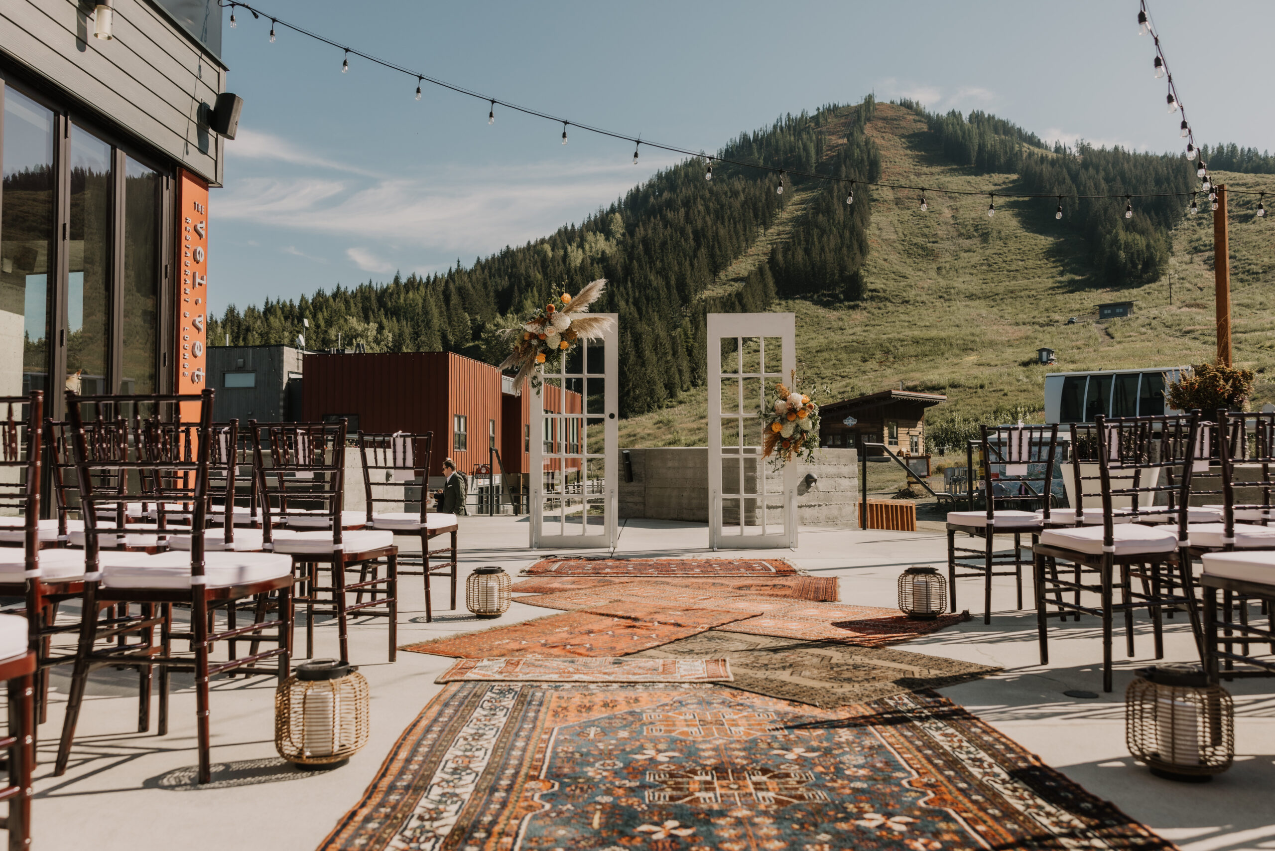 An outdoor wedding ceremony setup on a spacious terrace with a mountain backdrop. The aisle is lined with patterned vintage rugs and decorated with lanterns, leading up to a unique ceremony arch made from two tall white doors adorned with floral arrangements. Wooden chairs are neatly arranged on either side of the aisle, and string lights are hung overhead, adding a warm, inviting touch to the mountain-view setting.