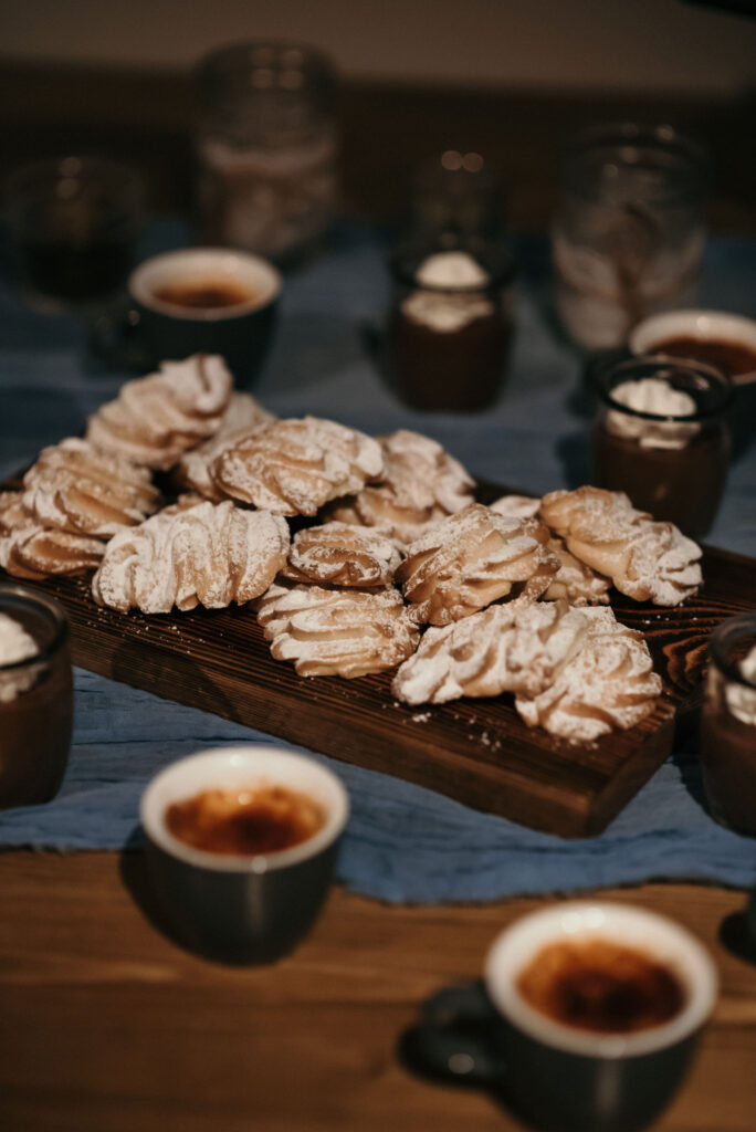 Delicious dessert spread at a wedding reception featuring a wooden board piled high with freshly baked, powdered sugar-dusted pastries, accompanied by small cups of espresso and chocolate mousse. The setup, laid out on a navy blue tablecloth, creates a warm and inviting coffee and dessert station for guests to enjoy.