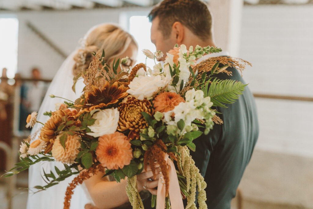 Close-up view of a bride and groom embracing, with a focus on a vibrant wedding bouquet held by the bride. The bouquet features a rich array of autumnal hues with sunflowers, dahlias, and delicate green ferns, embodying a rustic and warm wedding theme.