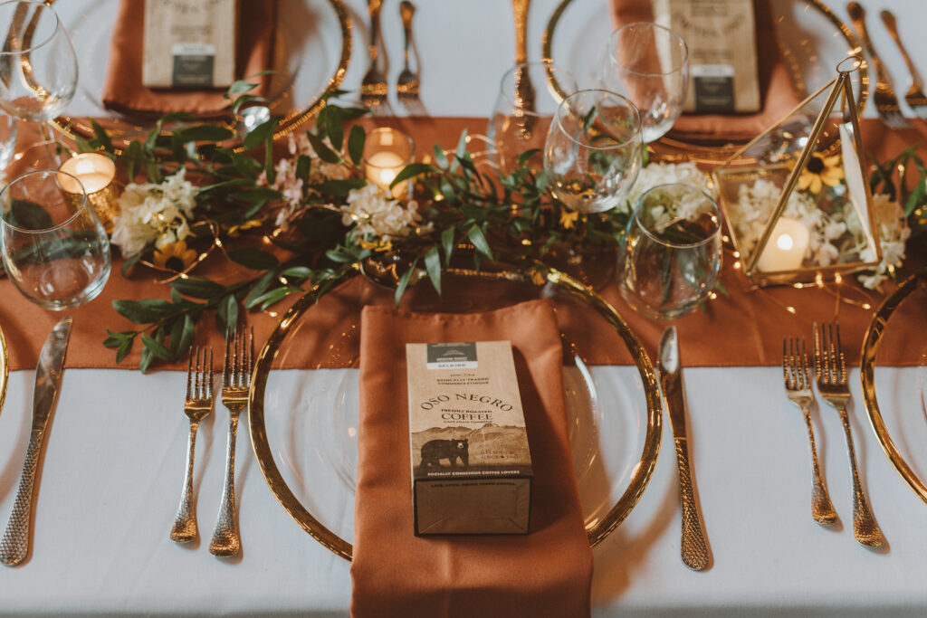 Elegant wedding table setting featuring a mocha satin table runner and gold flatware. Each setting includes a personalized coffee favor bag, surrounded by lush greenery and white florals, creating a warm and inviting ambiance for a sophisticated celebration.