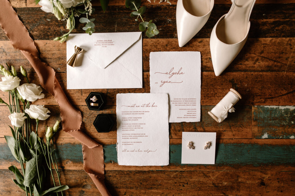 Flat lay of wedding details styled on a rustic wooden table, featuring handwritten invitations with deckled edges, a velvet ring box with a rose gold engagement ring, white heels, pearl earrings, a soft brown ribbon, and fresh white lisianthus blooms with greenery. The arrangement blends earthy tones with classic elegance, creating a timeless and romantic look.