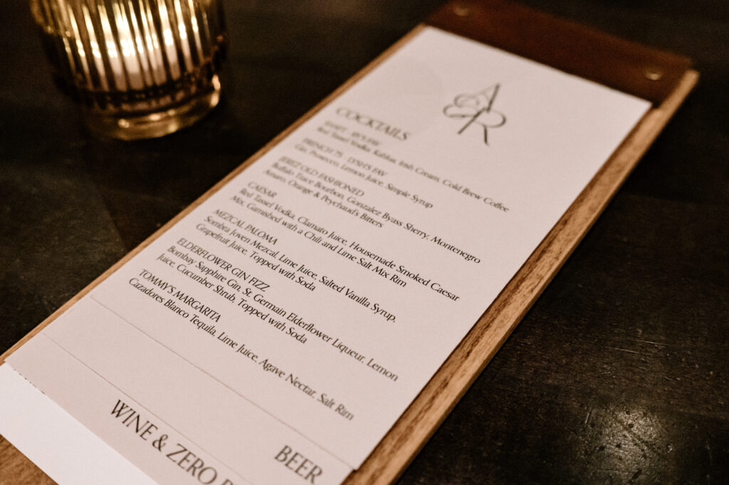 Elegant cocktail menu displayed on a wooden clipboard, featuring a sophisticated selection of signature drinks, surrounded by warm ambient lighting and a golden candle holder.