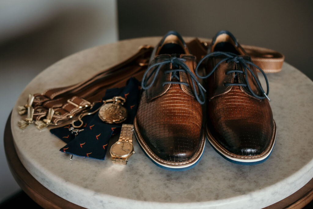 Elegant groom's accessories arranged on a marble table, featuring polished brown leather shoes with blue laces, a navy tie with fox motifs, gold cufflinks, a watch, and a leather belt, capturing the refined details of a wedding day ensemble.