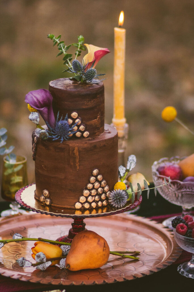 Artistic autumn wedding cake on a vintage plate, adorned with unique botanical elements including deep purple calla lilies, blue thistles, and an arrangement of hazelnut shells. The cake is complemented by a rustic candle and a display of fresh fruits and berries, set against an outdoor backdrop, creating an enchanting and cozy fall celebration scene.