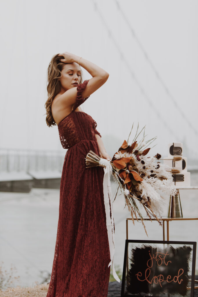 Stunning bride in a romantic deep burgundy lace dress holding a bohemian bridal bouquet, with a backdrop of a misty bridge. The bouquet features earthy tones with dried florals and feathers, complementing her elegant and moody elopement setting.