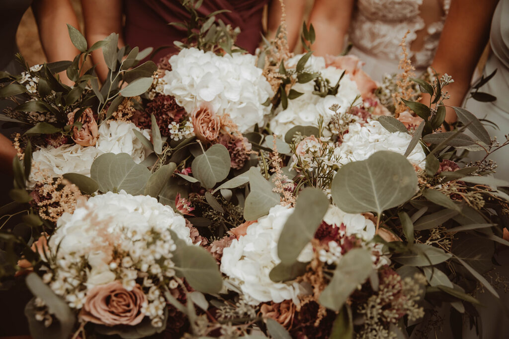 Close-up of bridesmaids' bouquets featuring a lush mix of white hydrangeas, blush roses, deep burgundy blooms, and abundant eucalyptus greenery. The floral arrangements are rich in texture and color, beautifully contrasting against the bridesmaids' dresses, perfectly capturing the essence of an elegant and romantic wedding style.