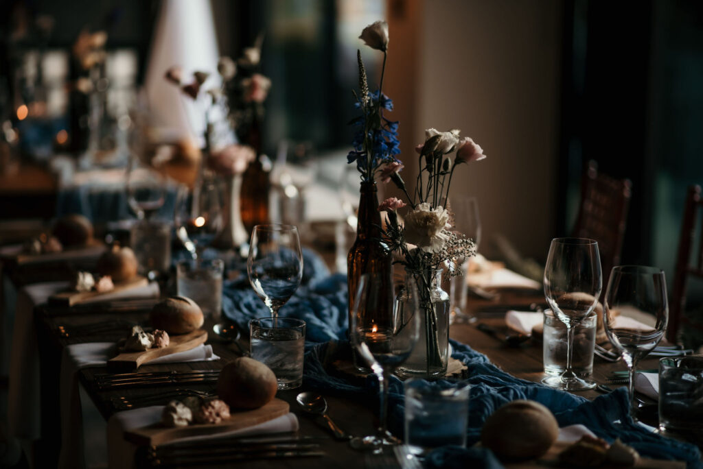 Moody and atmospheric wedding table setting with a rustic charm, featuring blue and brown tones. The table is adorned with clear glass bottles filled with blue delphiniums and pale pink roses, complemented by soft candlelight and simple bread rolls on wooden cutting boards, creating an intimate dining experience.