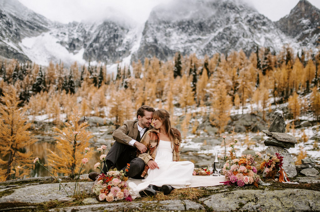 Romantic mountain elopement featuring a couple seated on a rocky outcrop surrounded by golden larch trees and snow-dusted peaks. The bride wears a white dress and cozy jacket, while the groom complements her in a brown suit. A vibrant floral arrangement with blush, burgundy, and peach hues adds warmth to the intimate outdoor setting.