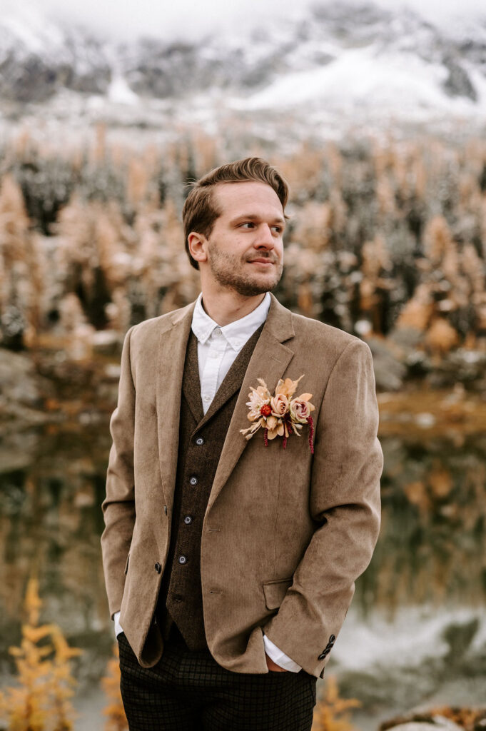 Groom in a stylish autumn wedding outfit, wearing a brown tweed suit with a white shirt and a brown vest, complemented by a floral boutonniere. He stands against a stunning backdrop of golden larch trees and snow-capped mountains, reflecting the natural beauty of a mountain wedding.