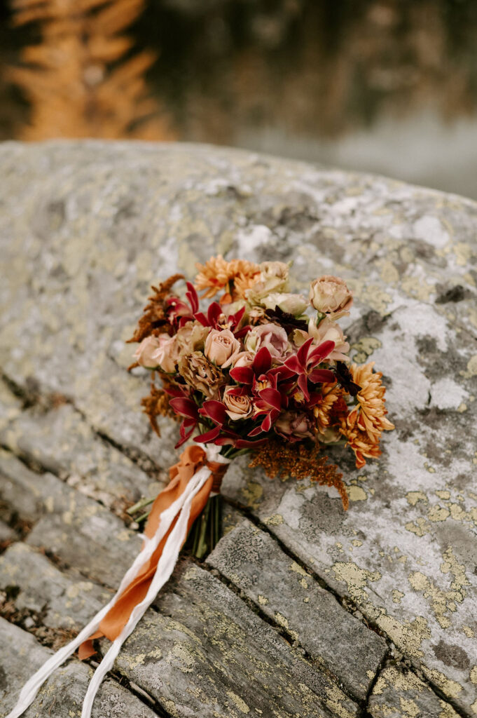 Artfully arranged bridal bouquet resting on a rugged stone surface, featuring a blend of autumnal tones with deep red leucadendron, peach roses, and accents of golden mums. The bouquet is tied with long, flowing ribbons in shades of cream and burnt orange, complementing the natural, earthy backdrop.