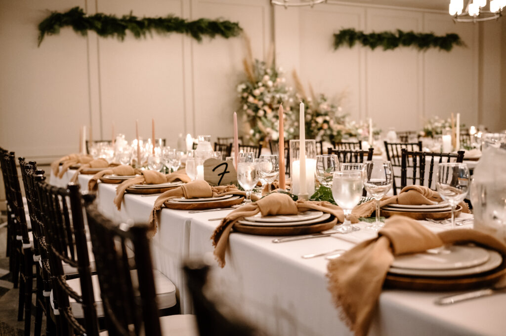 Elegant wedding reception setup featuring long tables with white linens and caramel-colored napkins neatly tied on dinner plates. The table decor includes tall white candles, crystal clear glassware, and lush greenery running the length of the table, framed by classic black chairs, creating a sophisticated and inviting atmosphere.