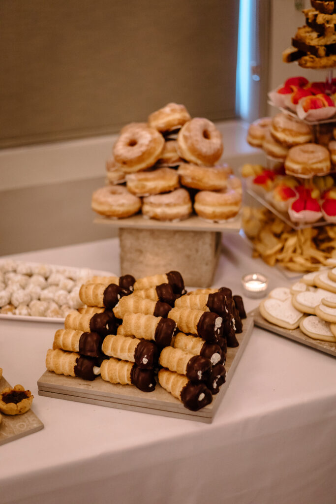 Delightful dessert buffet at a wedding featuring stacks of chocolate-dipped cream-filled pastries, a tower of glazed donuts, and a variety of other sweet treats on elegant serving trays, beautifully arranged to entice guests with a tempting selection of confections.