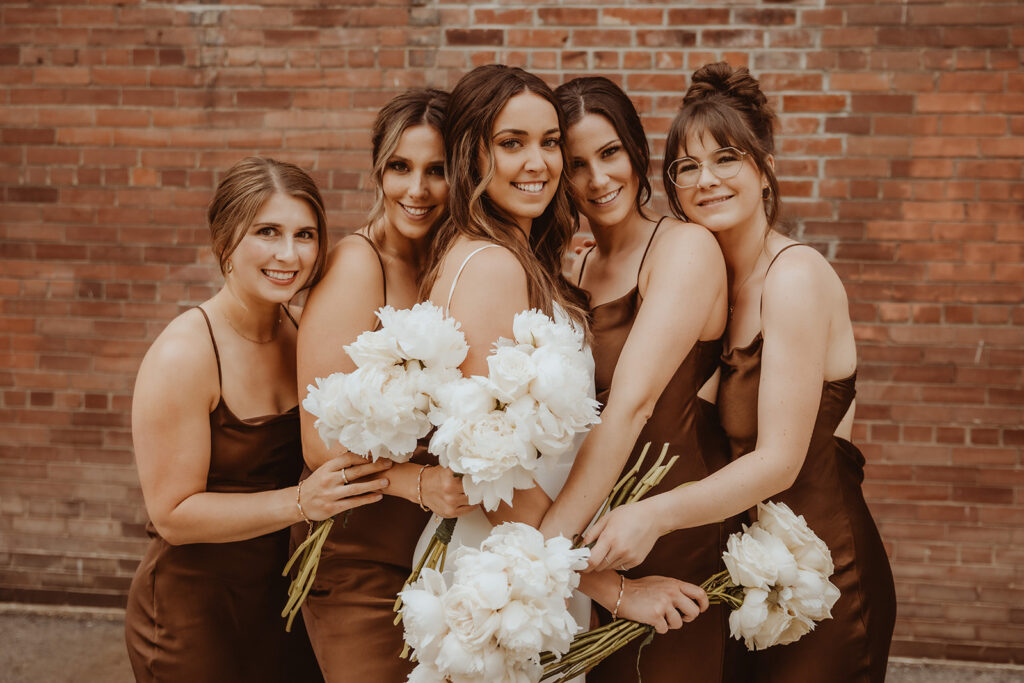 Radiant bride surrounded by her bridesmaids in rich mocha satin dresses, each holding lush white peony bouquets, against a warm brick wall backdrop.