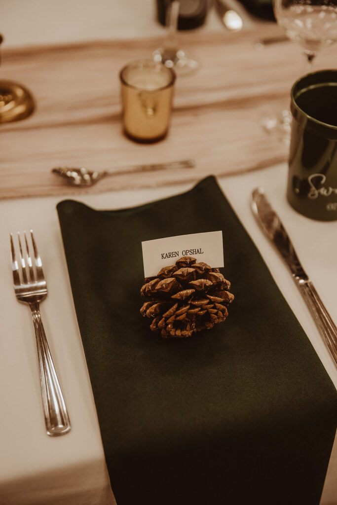 Elegant wedding place setting featuring a dark green napkin topped with a natural pine cone holding a place card. The setting is completed with classic silverware and golden candle holders, creating a cozy, rustic atmosphere at the reception table.