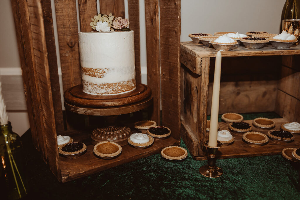 Rustic dessert table setup at a wedding, featuring a two-tiered cake with exposed sponge and cream frosting, decorated with subtle floral accents on a wooden stand. Surrounding the cake are various tarts with toppings like berries and meringue, displayed alongside a classic golden candlestick, all arranged on a reclaimed wooden table for a charmingly old-world feel.