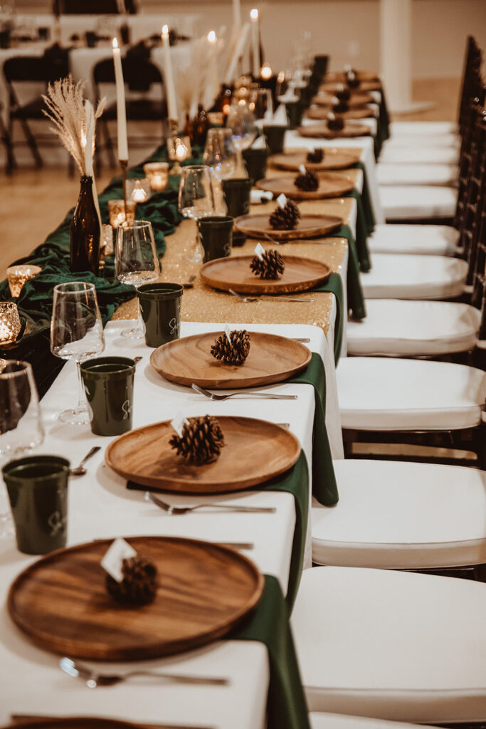 Rustic and elegant long wedding reception table setting with dark green table runners and golden sparkly tablecloths. The table features wooden charger plates, pine cone place card holders, and simple white candles, complemented by dark green mugs and glassware, creating a cozy, festive atmosphere.