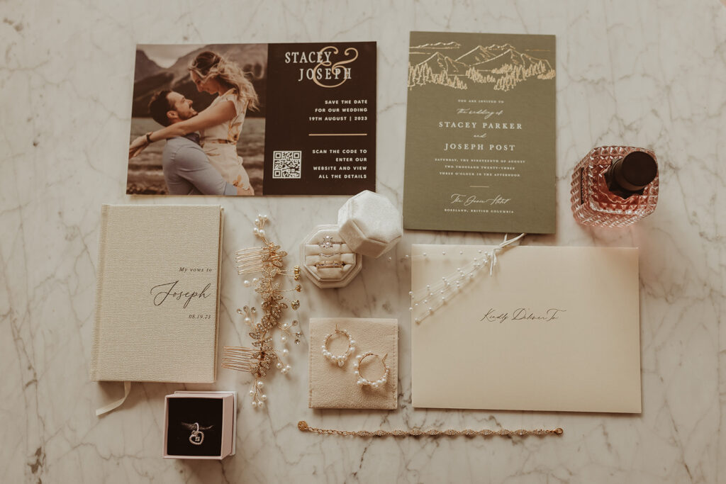 Flat lay of elegant wedding details displayed on a marble surface, including personalized vow books, gold and pearl jewelry, wedding invitations with mountain-inspired designs, a save-the-date card featuring the couple, and a velvet ring box. The arrangement exudes a timeless and romantic aesthetic with soft neutral tones and delicate accents.