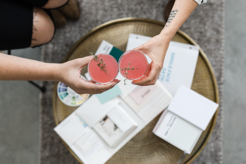 Two hands holding pink cocktails garnished with sprigs of thyme, toasting over a brass table filled with wedding stationery, color swatches, and planning materials.