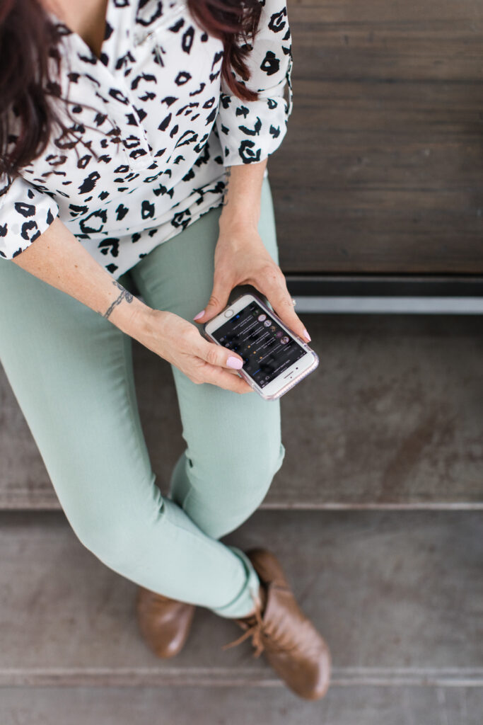 A stylishly dressed person in sage green pants and a black-and-white patterned blouse sits on a step while scrolling through their phone, possibly checking event details or vendor contacts.