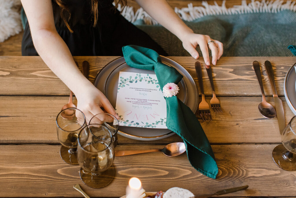 A close-up of a wedding reception place setting featuring a dark grey plate, emerald green knotted napkin with a floral accent, a printed menu card with botanical details, and gold-toned cutlery on a rustic wooden table.