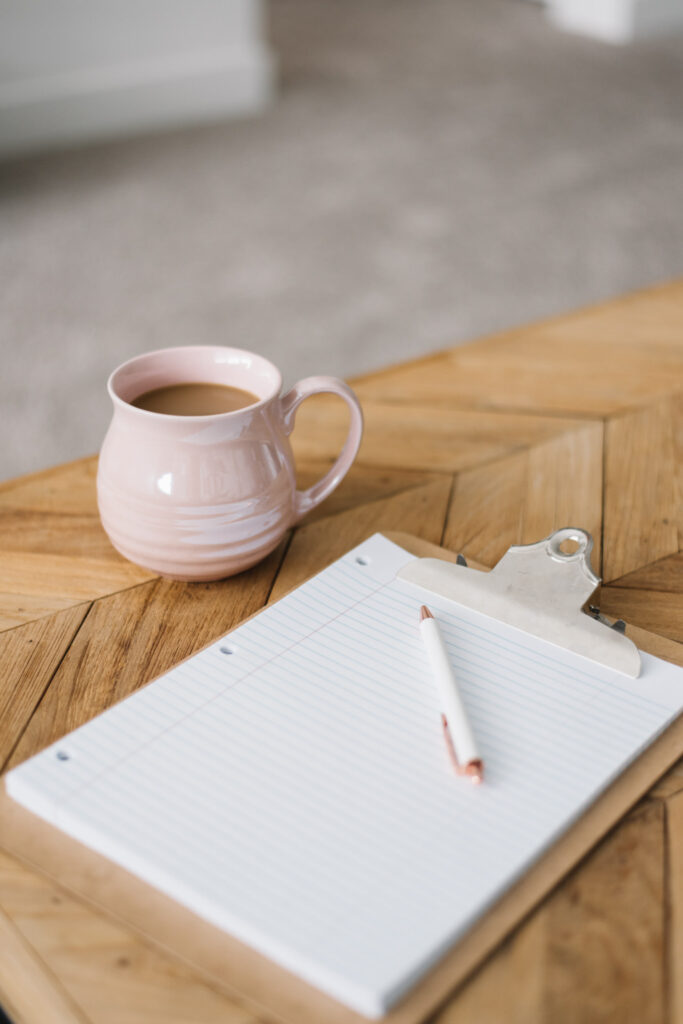 A cozy wedding planning setup with a pink ceramic mug filled with coffee next to a blank lined clipboard and a rose gold pen on a wooden table, ready for note-taking.