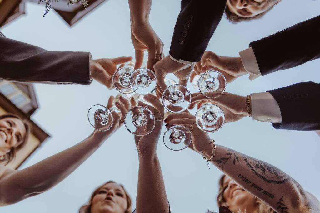 A dynamic low-angle shot of a group of people raising champagne glasses in a celebratory toast against a bright blue sky, showcasing hands adorned with rings and bracelets.